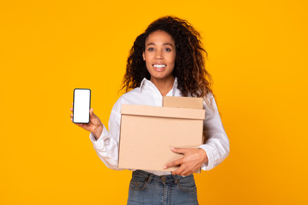 Black Woman Showing Smartphone Screen Holding Carton Box, Yellow Backgroundの写真素材