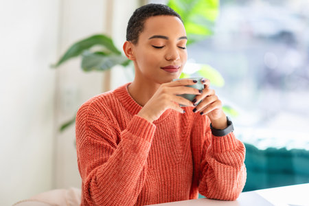 Relaxed young woman enjoys the aroma of her drink, eyes closed, wearing a cozy orange sweaterの写真素材