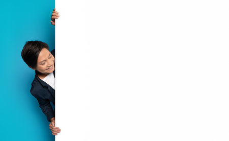 Asian man peeks playfully around a blank white vertical banner with a big smileの写真素材
