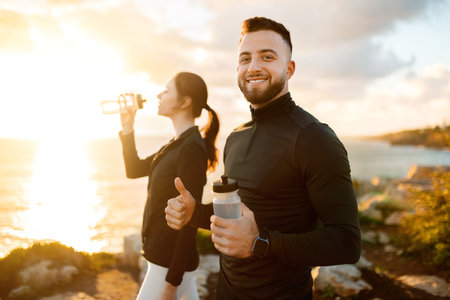 Active couple on coastal trail, man with water bottle giving thumbs up, woman drinking in backgroundの写真素材