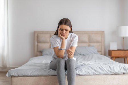 Sad disappointment lady holding pregnancy test, sitting on bed alone in bedroom interiorの写真素材