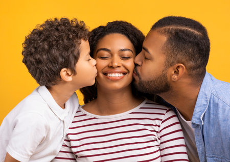 Adorable son and husband giving kisses on cheeks of smiling black motherの写真素材