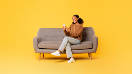 Black teen girl browsing on smartphone on couch, yellow backdropの写真素材