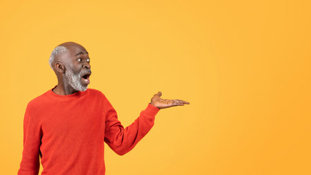 Astonished senior man in vibrant red sweater presents something on his open handの写真素材