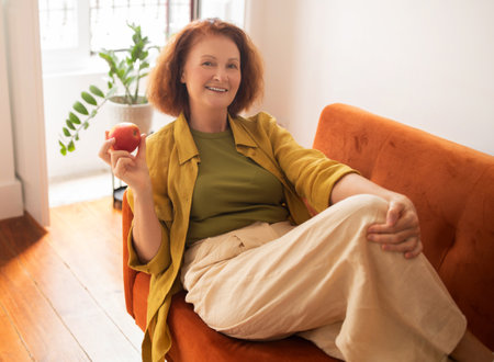 Smiling senior woman holding red apple and relaxing on couch at homeの写真素材