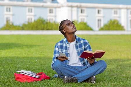 Thoughtful black guy reading book in park and using smartphone