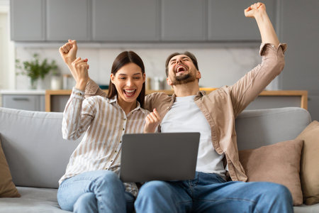 Excited caucasian couple with laptop celebrating success at homeの写真素材