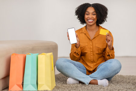 Positive millennial african american curly lady sit on floor with bags, show credit cardの写真素材