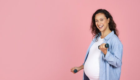 Smiling pregnant brunette lady exercising with barbells on pink backgroundの写真素材