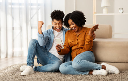 Glad excited young black couple winners, with smartphone celebrate win, success, rise fist, sit on...