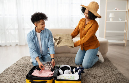 A smiling couple is packing for a trip, with the woman handing clothes to the man who is organizing...