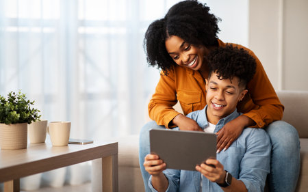 Happy young african american man and lady, hugs, use tablet, watch video on deviceの写真素材