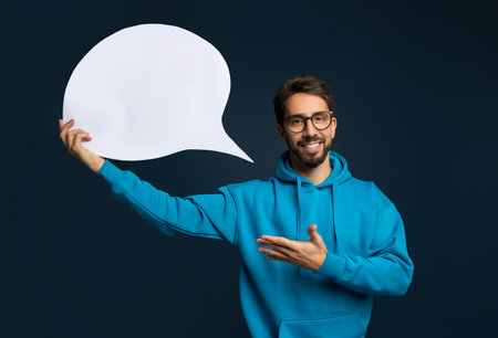 Cheerful young man in eyeglasses and blue hoodie holding blank speech bubbleの写真素材