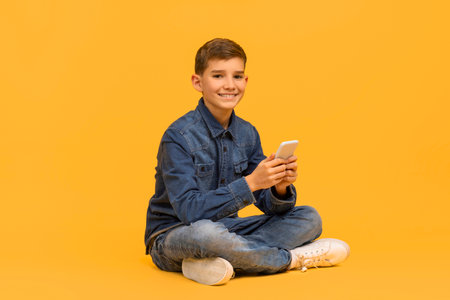 Smiling teen boy with smartphone in hands sitting cross-legged on yellow backgroundの写真素材