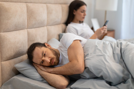 Man lying in bed with concern while woman uses phoneの写真素材