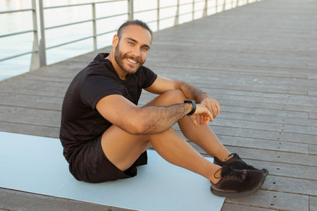 Happy sporty guy takes break sitting on gym mat outside