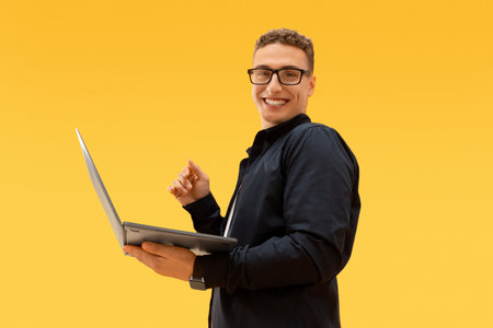 Young guy wearing eyeglasses holding laptop on yellow backgroundの写真素材