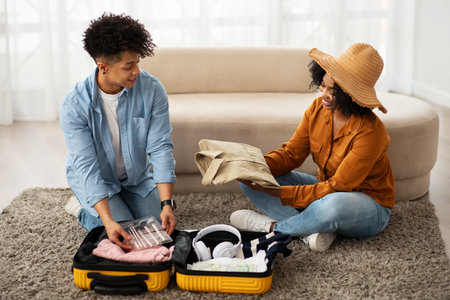 Engaged African American couple sitting on the floor, joyfully packing clothing into a bright yellow...
