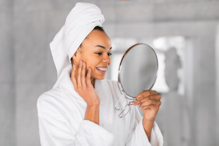 Pretty black woman holds mirror gently touching face in bathroomの写真素材