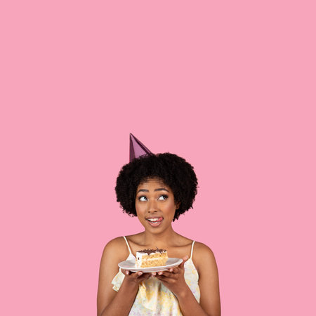 An astonished young woman in a party hat, holding a slice of cake on a plate, looks up with wide eyesの写真素材
