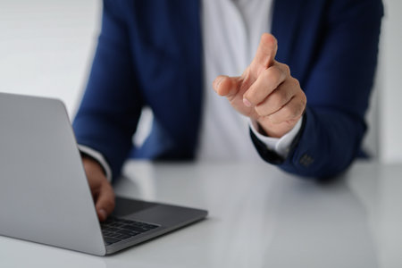 A businessman in a blue suit is pointing towards the camera while working on his laptop at a white deskの写真素材