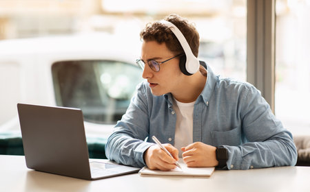 A young, curly-haired man wearing headphones writes in a notebook while working