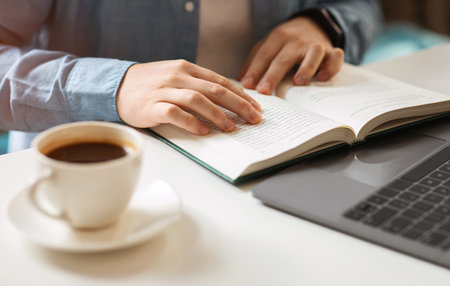 Close-up of a persons hands reading a book with a cup of coffee on the side and a laptop in the...