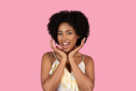 An exuberant young African American woman with curly afro hair joyfully holds her faceの写真素材