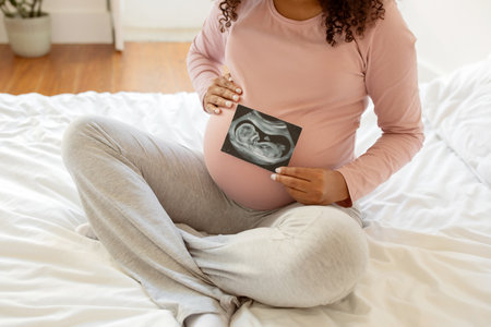 Unrecognizable black pregnant woman showing baby sonography photo while sitting on bed