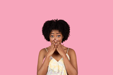 A surprised young African American woman with a natural afro hairstyle covers her mouth with her...