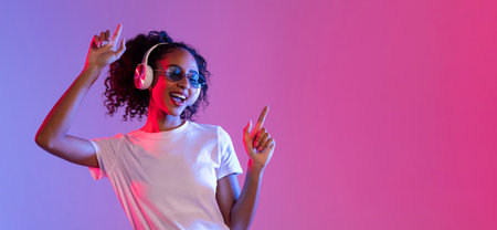 Joyful black woman with sunglasses and headphones dancing on neon background, free spaceの写真素材