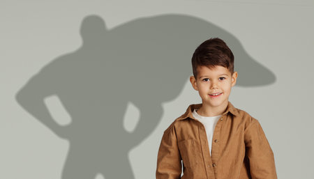 A happy young boy in a brown jacket smiles brightly, standing in front of a gray backdrop with a...