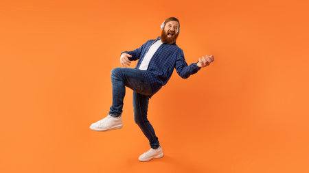 Energetic guy enjoying music through headphones playing invisible guitar, studio