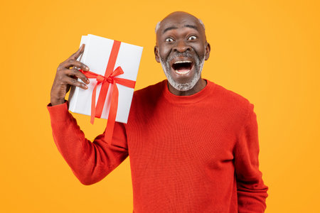 Ecstatic senior black man in red sweater holding a gift with a red ribbon to his earの写真素材