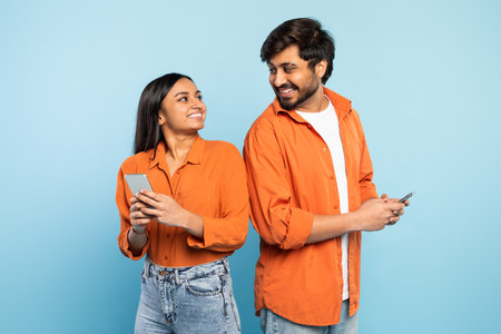 Excited hindu couple standing back to back using smartphones, studio