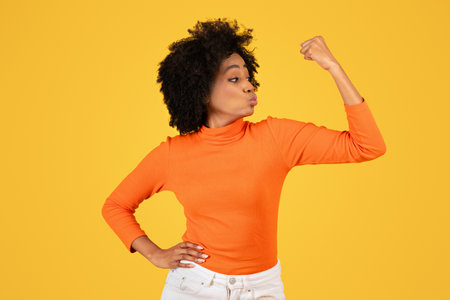 Confident young woman with afro hair flexing her arm muscle in a strong pose, wearing an orange turtleneckの写真素材