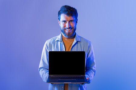 Smiling Young Man Holding Laptop in Front of Blue Backdrop