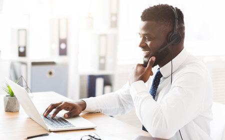 Cheerful young worker with headset sitting in front of laptop