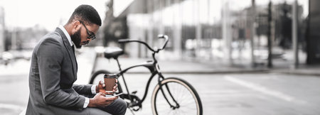 Businessman holding coffee cup and phone, sitting near bicycle outdoorsの写真素材