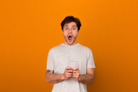 Man with shocked expression holding phone on orangeの写真素材