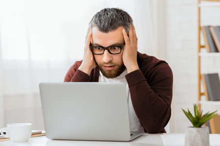 Man in Home Office Stares in Shock at Laptop Screen