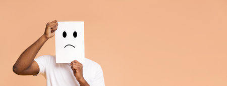 Man Holding Sad Face Sign in Front of Peach Backgroundの写真素材