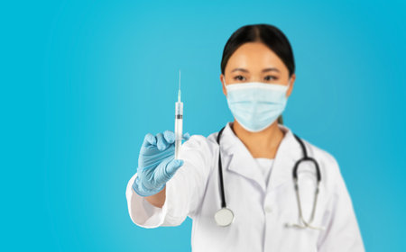 Woman Doctor Wearing Face Mask Holds Syringe Over Blue Backgroundの写真素材