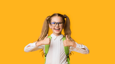 Cheerful Little Schoolgirl Gesturing Thumbs-Up With Both Hands, Yellow Background