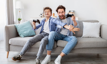 Joyful dad and son with joysticks playing video games at home, boy distracting father with hand