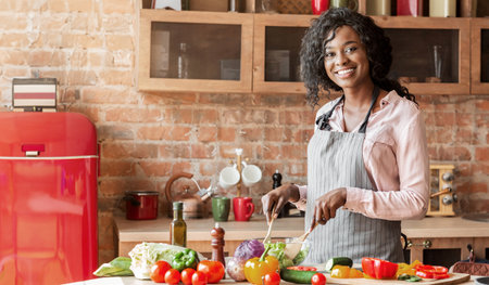 Joyful african lady mixing veggies in bowl in kitchenの写真素材