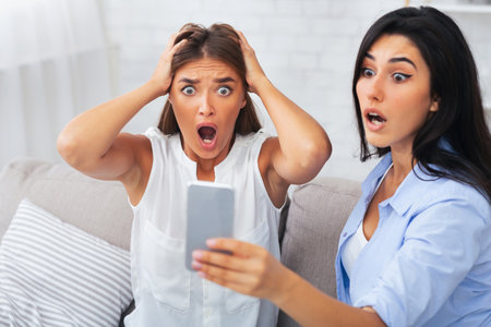 Shocked Girls Looking At Smartphone Screen Sitting On Couch Indoorの写真素材