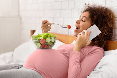 Young expectant eating vegetarian salad and talking on phoneの写真素材