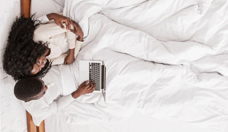 Afro couple sitting in bed with laptop, top view