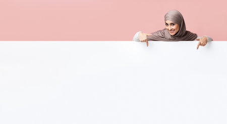 Cheerful arabic girl in headscarf pointing at white blank advertisement boardの写真素材
